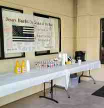 A table in the church courtyard containing orange juice, water and coffee.  Above the table hangs a banner signed by supporters.  The banner states "Jesus Backs the Blue and Green".
